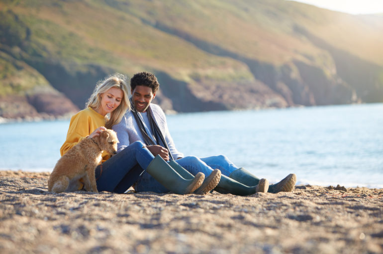 Loving Couple Sitting On Sand As They Walk With Dog Along Shoreline On Winter Beach Vacation