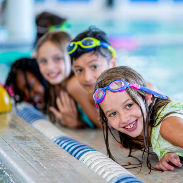 A multi-ethnic group of kids are indoors in a pool. Some of them are wearing goggles and smiling at the camera.