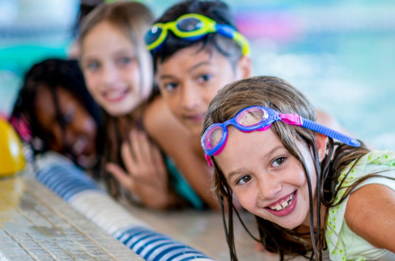 A multi-ethnic group of kids are indoors in a pool. Some of them are wearing goggles and smiling at the camera.