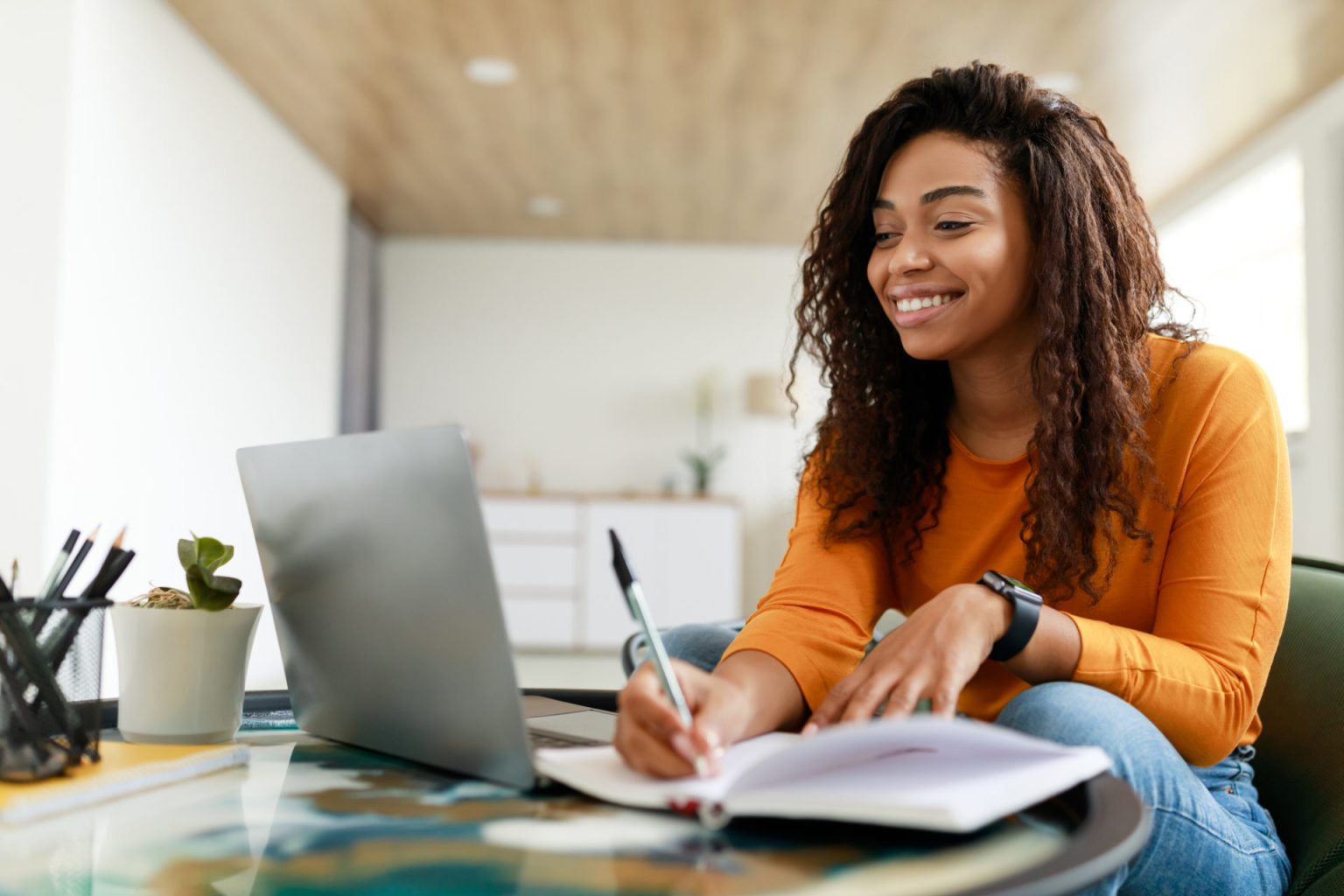 Smiling young African American woman sitting at desk working on laptop and writing letter in paper notebook, holding pen and looking at screen.